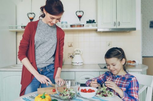 Happy mother and cheerful daughter cook salad together in the kitchen at home cutting vegetables and talking. Family, cooking, and people concept